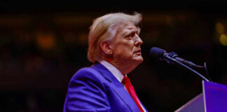 A man in a blue suit and red tie speaking into a microphone on stage