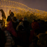 A crowd of migrants gathered near a barbed wire fence at night