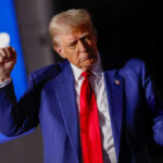 A man in a blue suit and red tie gesturing during a speech at a rally