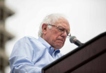 A man with white hair and glasses speaking into a microphone at an outdoor event