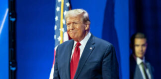 A man in a suit smiling at a political event with an American flag in the background