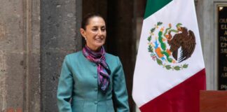 A woman in formal attire stands next to the Mexican flag, smiling at an official event