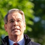 Treasury Nominee DEFENDS Trump’s Outrageous Comment A man in formal attire speaking outdoors with a blurred green background