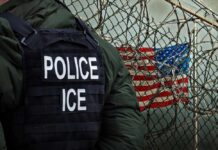 A police officer in a tactical vest stands near a barbed wire fence with an American flag in the background