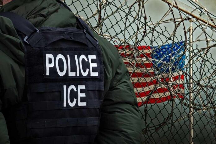 A police officer in a tactical vest stands near a barbed wire fence with an American flag in the background