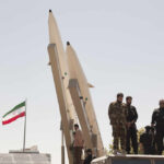Military personnel standing near missile launchers with an Iranian flag in the background