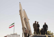 Military personnel standing near missile launchers with an Iranian flag in the background