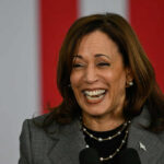 A woman smiling while speaking at a podium with an American flag backdrop