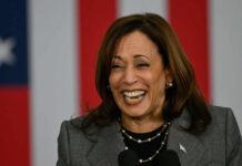 A woman smiling while speaking at a podium with an American flag backdrop