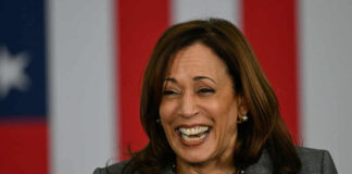 A woman smiling while speaking at a podium with an American flag backdrop