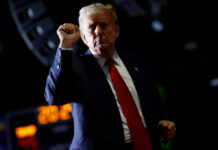 A man in a suit with a red tie raises his fist in a confident gesture against a dark background