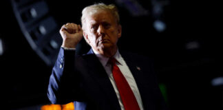 A man in a suit with a red tie raises his fist in a confident gesture against a dark background
