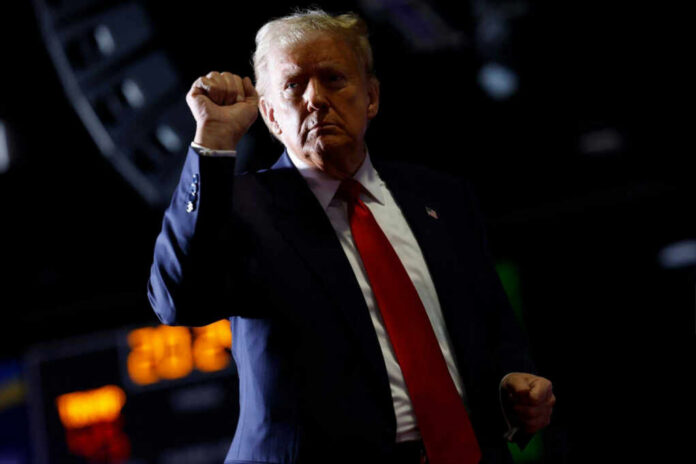 A man in a suit with a red tie raises his fist in a confident gesture against a dark background
