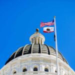 View of a state capitol dome with American and California flags against a blue sky