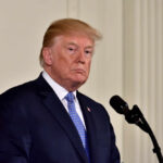 A political figure standing at a microphone with a serious expression