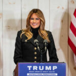 A woman in a black jacket smiling at a podium with a Trump sign and American flags in the background