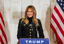 A woman in a black jacket smiling at a podium with a Trump sign and American flags in the background