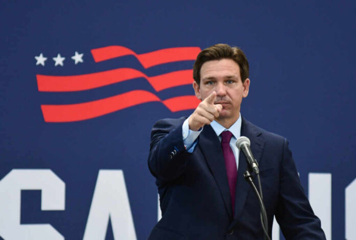 A man in a suit pointing while speaking into a microphone against a backdrop featuring an American flag design