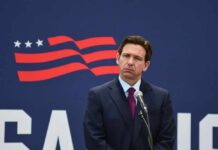 A man in a suit standing at a microphone with a serious expression against a backdrop featuring a stylized American flag