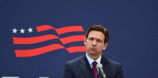 A man in a suit standing at a microphone with a serious expression against a backdrop featuring a stylized American flag