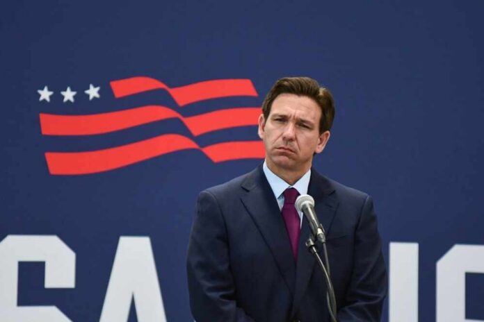 A man in a suit standing at a microphone with a serious expression against a backdrop featuring a stylized American flag