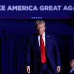 A man in a suit with a red tie walking towards the camera at a political rally