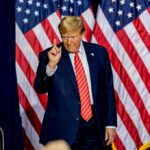 A man in a suit gesturing with one finger in front of multiple American flags