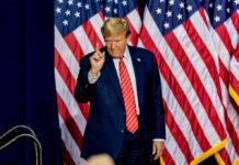 A man in a suit gesturing with one finger in front of multiple American flags