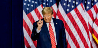 A man in a suit gesturing with one finger in front of multiple American flags