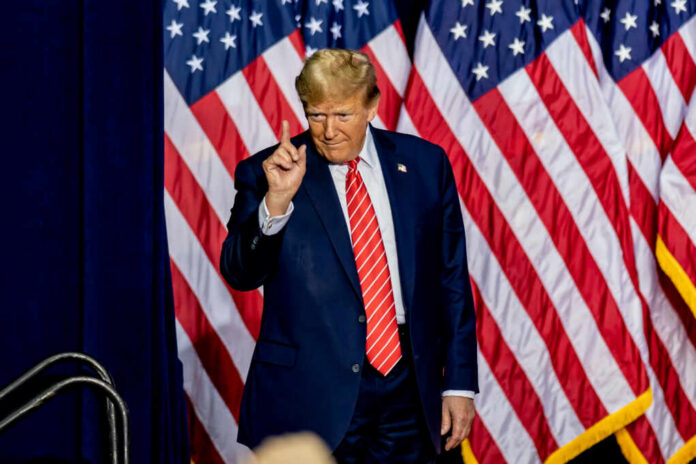 A man in a suit gesturing with one finger in front of multiple American flags