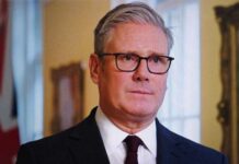 A man in formal attire with glasses, standing indoors with a serious expression