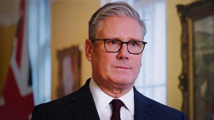 A man in formal attire with glasses, standing indoors with a serious expression