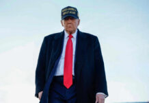 A political figure wearing a black coat and red tie, with a Make America Great Again hat, walking outdoors
