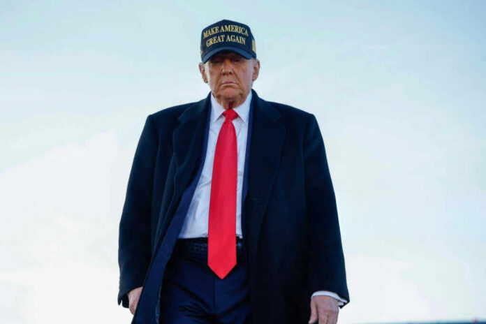 A political figure wearing a black coat and red tie, with a Make America Great Again hat, walking outdoors