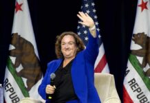 Woman in blue suit waving while seated with microphone