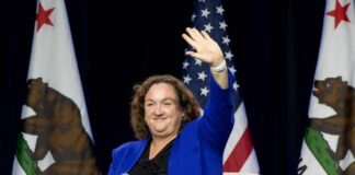 Woman in blue suit waving while seated with microphone