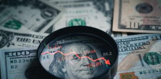 America’s Next Economic Test Looms Magnifying glass over US dollar bills with stock market data displayed