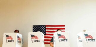 Individuals casting their votes in a polling station with an American flag in the background