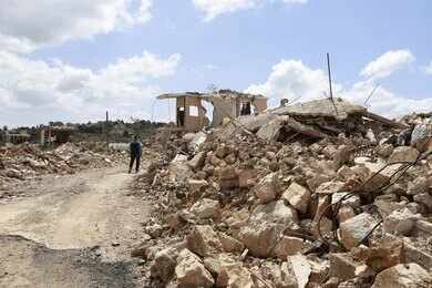 2645985195 Person walking past rubble and destroyed buildings on a dirt road