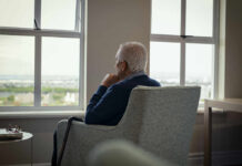 Elderly man sitting in a chair, looking out a window with a thoughtful expression