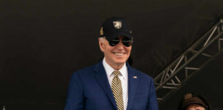 A smiling man in a suit and sunglasses at a military event, with a woman in uniform beside him
