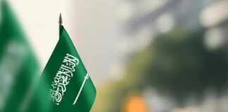 A green flag of Saudi Arabia with white Arabic text and a sword, set against a blurred background