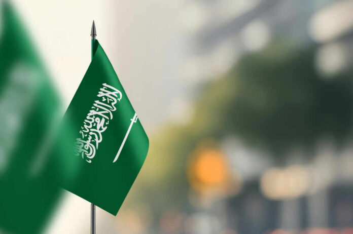 A green flag of Saudi Arabia with white Arabic text and a sword, set against a blurred background