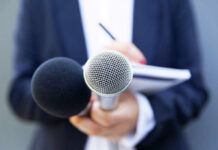 A person holding two microphones, one black and one silver, with a notebook in the background