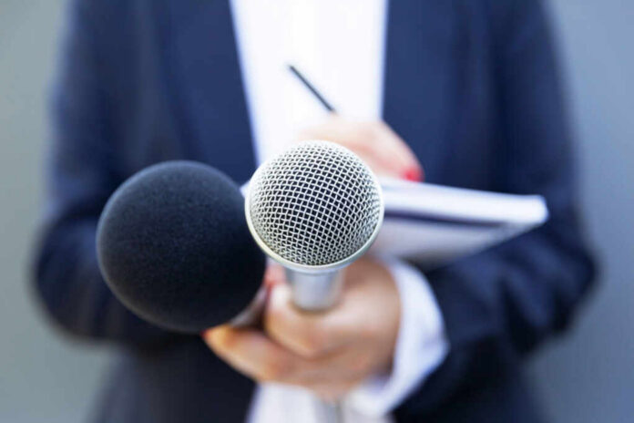 A person holding two microphones, one black and one silver, with a notebook in the background