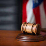 A wooden gavel resting on a round base with a blurred American flag in the background