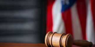 A wooden gavel resting on a round base with a blurred American flag in the background