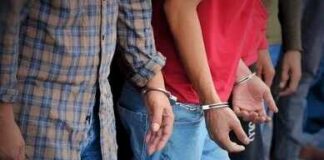 Identity Theft Ring Exposed at Window Plant A police officer in a tactical vest stands near a barbed wire fence with an American flag in the background