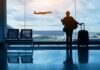 Silhouette of a traveler holding a coffee cup and standing next to a suitcase, looking out at an airplane taking off from the airport