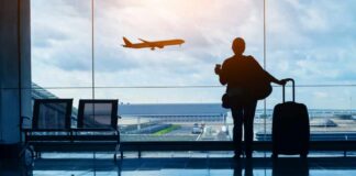 Silhouette of a traveler holding a coffee cup and standing next to a suitcase, looking out at an airplane taking off from the airport